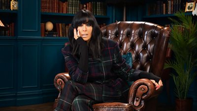 Claudia Winkleman sits in a high backed chair with her legs crossed, resting her face on her hand. She wears a dark black long sleeve top and tartan trousers.