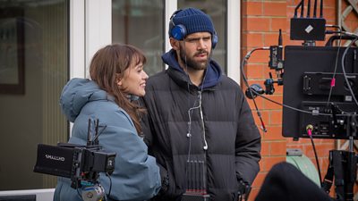 A man and a woman stand outdoors on set together in warm coats, watching something on a monitor