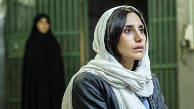 A woman in a white headscarf sits at a table in a prison looking worried. A blurry figure in black is visible behind her.