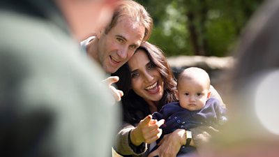 A man and woman smile holding a baby and pointing towards a camera