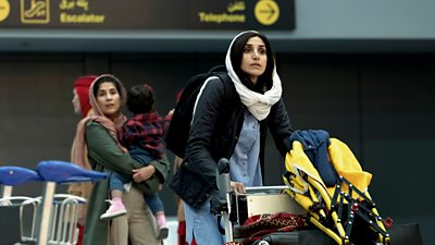 Two women walk through an airport, one pushing a trolley of luggage and the other holding a baby