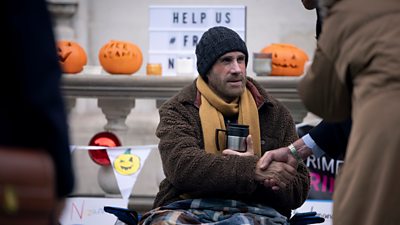A man in a hat, scarf and coat sits on the ground outdoors, surrounded by pumpkins and Halloween banners as her stages a protest
