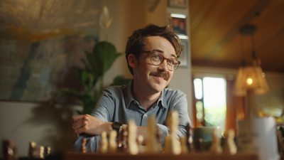 A young man sat in front of a chess board with a pot plant behind him. He is smiling.