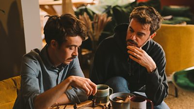 Two young men - one is pondering his next chess move over a cup of tea and a biscuit. They look thoughtful.
