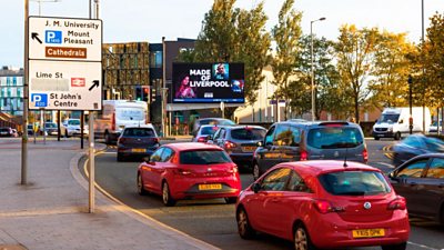 Made in Liverpool artwork on a billboard with cars driving towards it on a busy street. A street sign and some trees are also in shot.