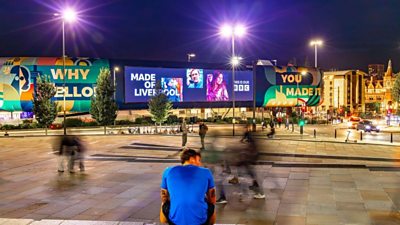 Made in Liverpool artwork on a large media wall in the city centre. It is night time and the art work is lit up. The people in shot are blurred.