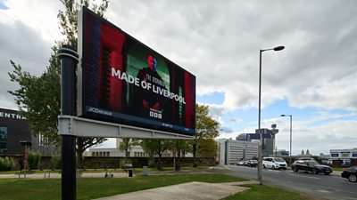 The Responder Made in Liverpool artwork on a large billboard against a grey and blue sky. There are parked cars, some buildings and trees also in shot.