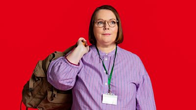Joanna Scanlan in character as Beth Thornton in Riot Women. Wearing a pink shirt, glasses, and a lanyard, she holds a backpack on her shoulder. 