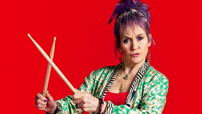 Lorraine Ashbourne in character as Jess Burchill in Riot Women. She wears a green and cream shirt and holds drum sticks together. Her hair is tied up and dyed purple. 