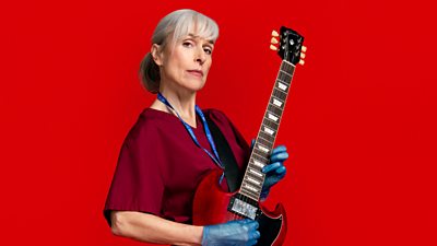 Amelia Bullmore in character as Yvonne Vaux in Riot Women. She wears red medical scrubs and blue gloves. She holds an electric guitar in both hands. 