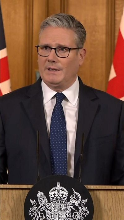Sir Keir Starmer in a black suit and navy blue tie, stood at a podium