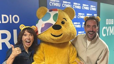 Pudsey stood in between Trystan and Emma, whoa re both wearing yellow Pudsey ears. They are stood in front of a Radio Cymru and Radio Cymru 2 branded wall.