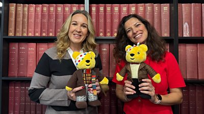Catrin and Behnaz holding Pudsey teddy bears dressed in brown jackets.  They are stood in front of old volumes of the Radio Times books with red spines.