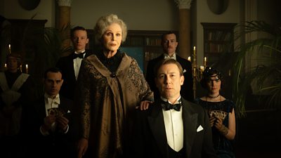 A group shot of six people dressed in old fashioned, smart clothing, as if at a party. The men are in tuxedos and the women in black dresses. One woman stands with her hand on the shoulder of a man at the front of the scene, which is dimly lit and eerie.