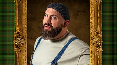 A man with a beard poses in dungarees, a light top and a beanie hat, looking to camera with a slightly questioning expression on his face.