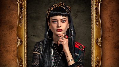A woman with long black hair looks to camera with a hand to her chin. She wears a leopard print headband and a jacket featuring multiple different patches of fabric along with silver hoop earrings. He image is set in a gilded frame.