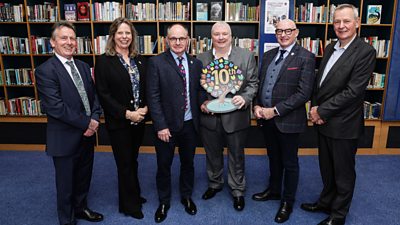 A group shot of six people stood on a blue carpet with book shelves behind them. They are smartly dressed and one of them is holding a Book Week trophy in the shape of a tree.