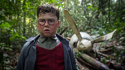 A boy wearing glasses is stood in a forest with a plane wreck behind him.  He is wearing a coat and jumper and his face is dirty.