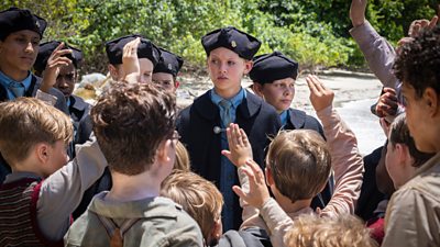 A group of slightly older children, wearing hats and uniforms are addressing a group of younger boys who have their hands in the air, as if they are wanting to answer a question or volunteer.