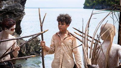 Three boys, one in a shirt and the other two topless but covered in body paint, holding long spears designed for fishing made out of sticks. They are next to the sea.