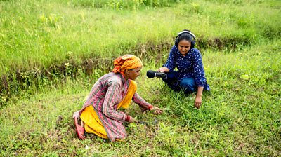 Bindu Chaudhary, journalist at work interviewing a woman farmer in the field. she ie wearing blue, the farm is wearing pink and yellow.