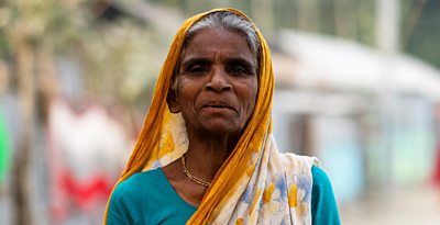 A women wearing turquoise and a yellow headscarf is staring in to the camera, she is in Bangladesh, she has grey hair and pensive expression.