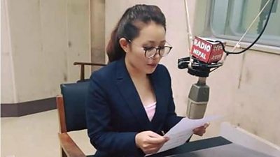 Women journalist Balika Maden wearing a navy blazer sitting at microphone at Radio Nepal station. The mic has a red 'Radio Nepal' sign on it. She is holding and reading from a piece paper