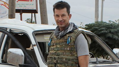 A man wearing a camouflage bullet-proof vest over a grey t-shirt smiles to the camera. He stands beside a pick-up truck by the side of a road.