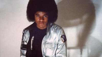 A photo of Michael Jackson wearing a silver jacket. He sits on a stool infant of a white wall and looks directly to the camera.