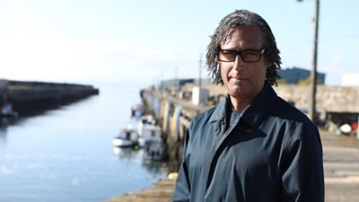 Professor David Olusoga stands beside a seaside dock. Small boats are moored in the background.