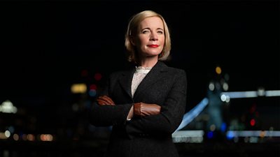 Lucy Worsley stands with her arms folded in front of a city skyline at night. Lights in the distance illuminate Tower Bridge in London.
