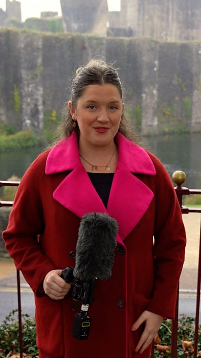 Menyw yn sefyll tu fas castell yn dal microffon ac yn gwisgo cot binc