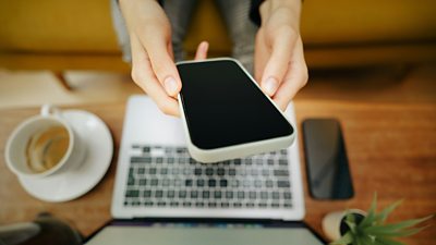 Hands holding a mobile phone above the keyboard of a laptop.