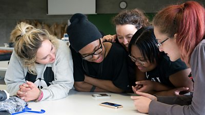A group of young people crowd over a desk where they are looking at mobile phones.