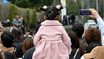 The back of a child who is sitting on someone's shoulders in a crowd of people. Some of the crowd are pointing mobile phones at something happening in the distance.