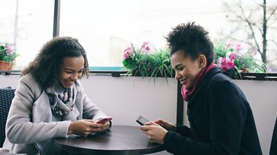 Two people sitting at a table, possibly in a coffee shop, who are smiling as they each look at their mobile phones.