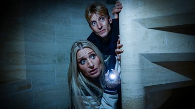 Daisy May and Charlie Cooper peer around a curved, stone staircase in a gloomy, stone building. Daisy May shines a torch at something in the distance.
