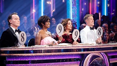 The Strictly Come Dancing judges sit behind a glittering desk, lifting their paddles to deliver scores of 8, 9, 9 and 8.