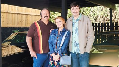 Two men with moustaches and loud shirts stood either side of a woman in a floral dress and denim jacket.  Two retro cars are in shot behind them.
