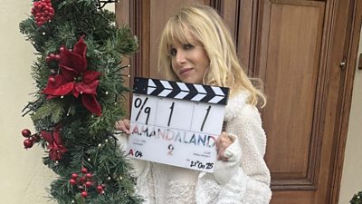 A woman in a white jacket stands outside a wooden door, decorated with festive greenery and red ribbon, holding a clapperboard for the start of filming on Amandaland