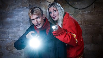 A man and woman peer into a dark space holding torches