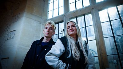 A man and woman stand looking down towards the camera against the backdrop of a towering window set in a stone wall