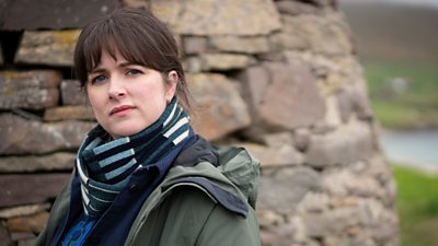 Alison O’Donnell in character as DI Alison ‘Tosh’ Mcintosh. She wears a coat and scarf and stands beside a large, stone monument. 