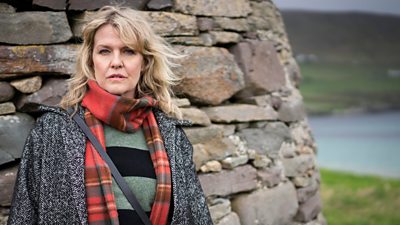 Ashley Jensen in character as DI Ruth Calder. She wears a warm coat and patterned scarf and stands beside a large, stone monument. 