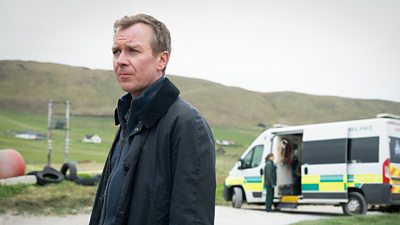 Steven Robertson in character as Sandy. He wears a coat and warm fleece and stands on tarmac beside hills and green countryside. An ambulance is parked in the background. 
