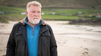 Lewis Howden in character as Sgt Billy McCabe. He stands on a sand beach. Green countryside and hills are visible in the background. 