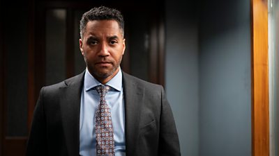 Samuel Anderson in character as Matt Blake. Wearing a suit, he stands in a darkened office and looks directly to the camera. 