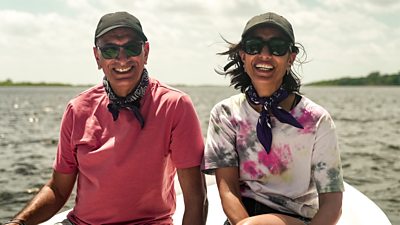 A man and a woman sit on a boat smiling. They wear matching scarves, baseball caps and sunglasses.