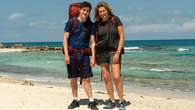 A man and woman stand together on a beach smiling as the sea laps at the shore behind them. They're wearing backpacks.