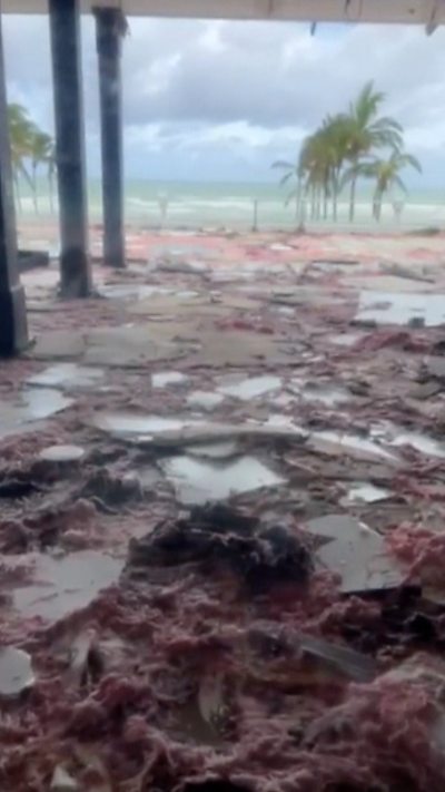 Debris of what appears to be panels and insulation fill a floor with the beach and palm trees in the background.
